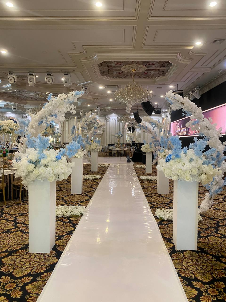 A ballroom aisle framed by tall arches of white and powder-blue blossom on white pedestals, above a white runner.