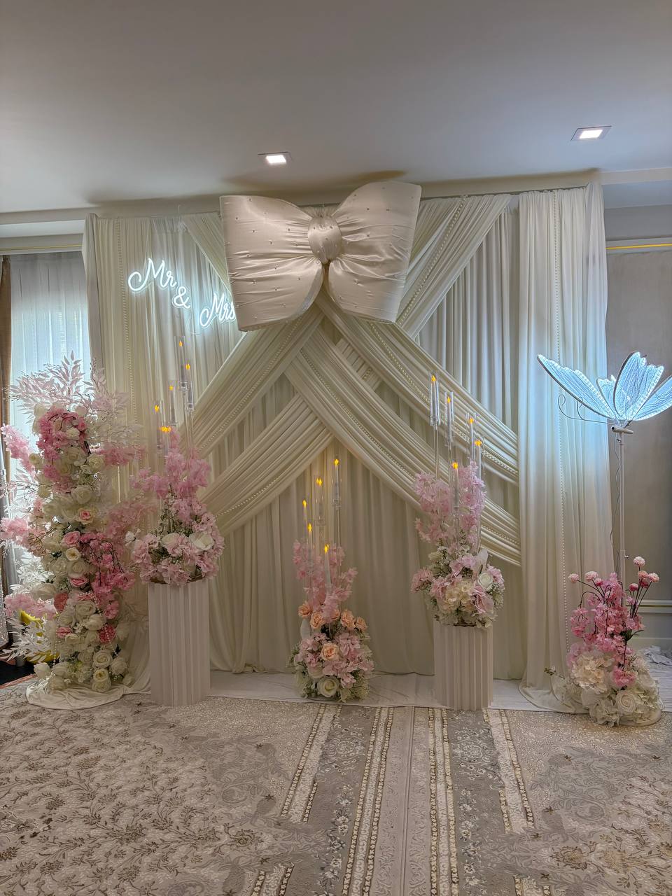 An ivory draped backdrop with an oversized pearl-studded satin bow, a Mr & Mrs neon sign and columns of blush roses.
