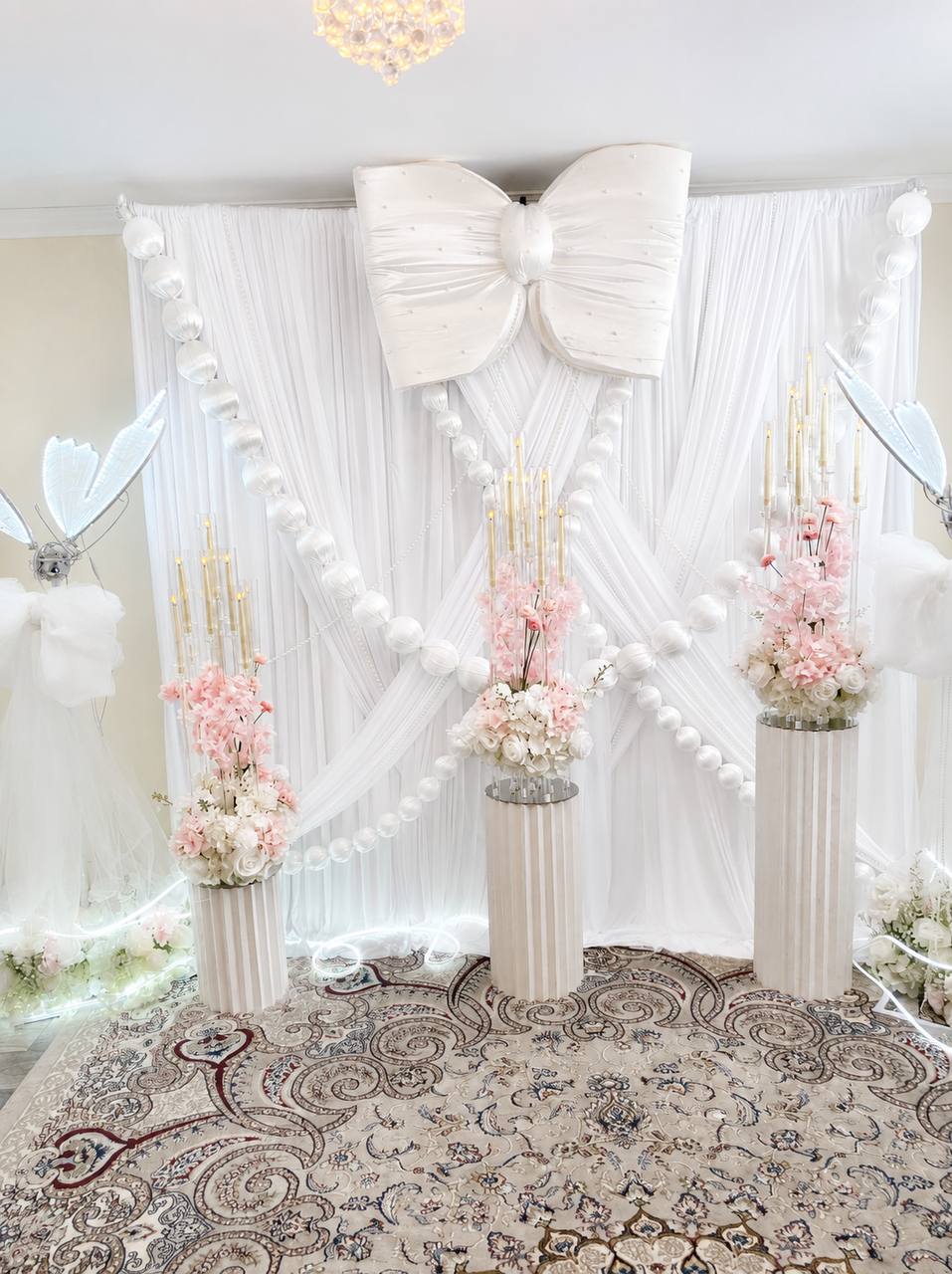 A white draped backdrop with a large satin bow and looping pearl garlands, framed by pedestals of blush blossom and gold candelabras.