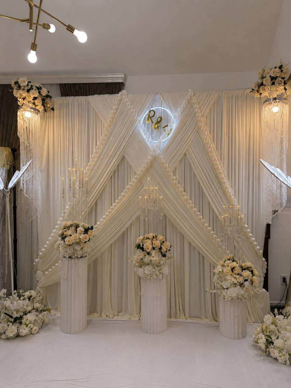 A cream pleated backdrop laced with oversized pearls and a lit monogram ring, with ivory rose pedestals and gold candelabras.
