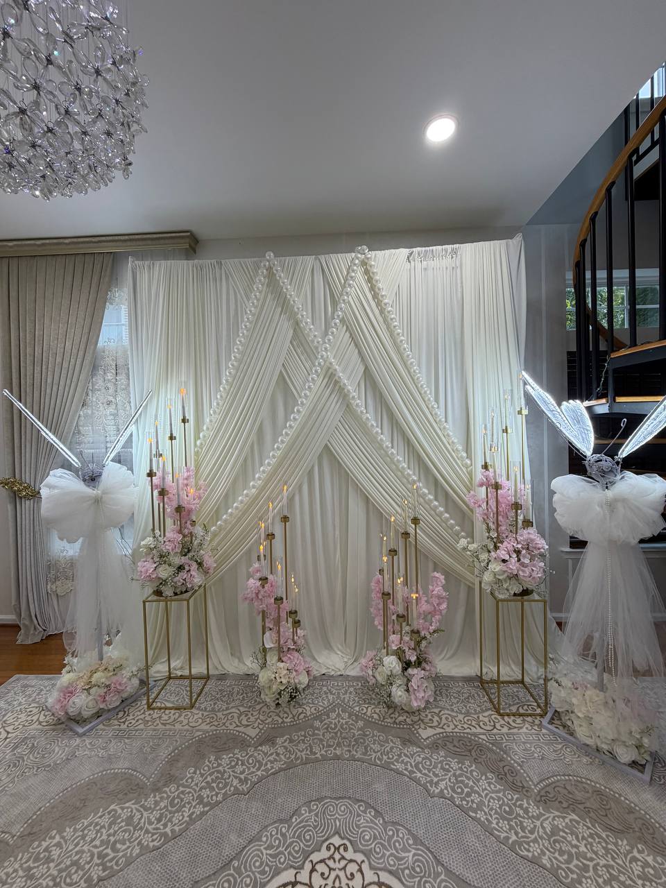 A crossed ivory drape backdrop trimmed with pearl garlands, flanked by lit angel-wing sculptures and blush floral pedestals.