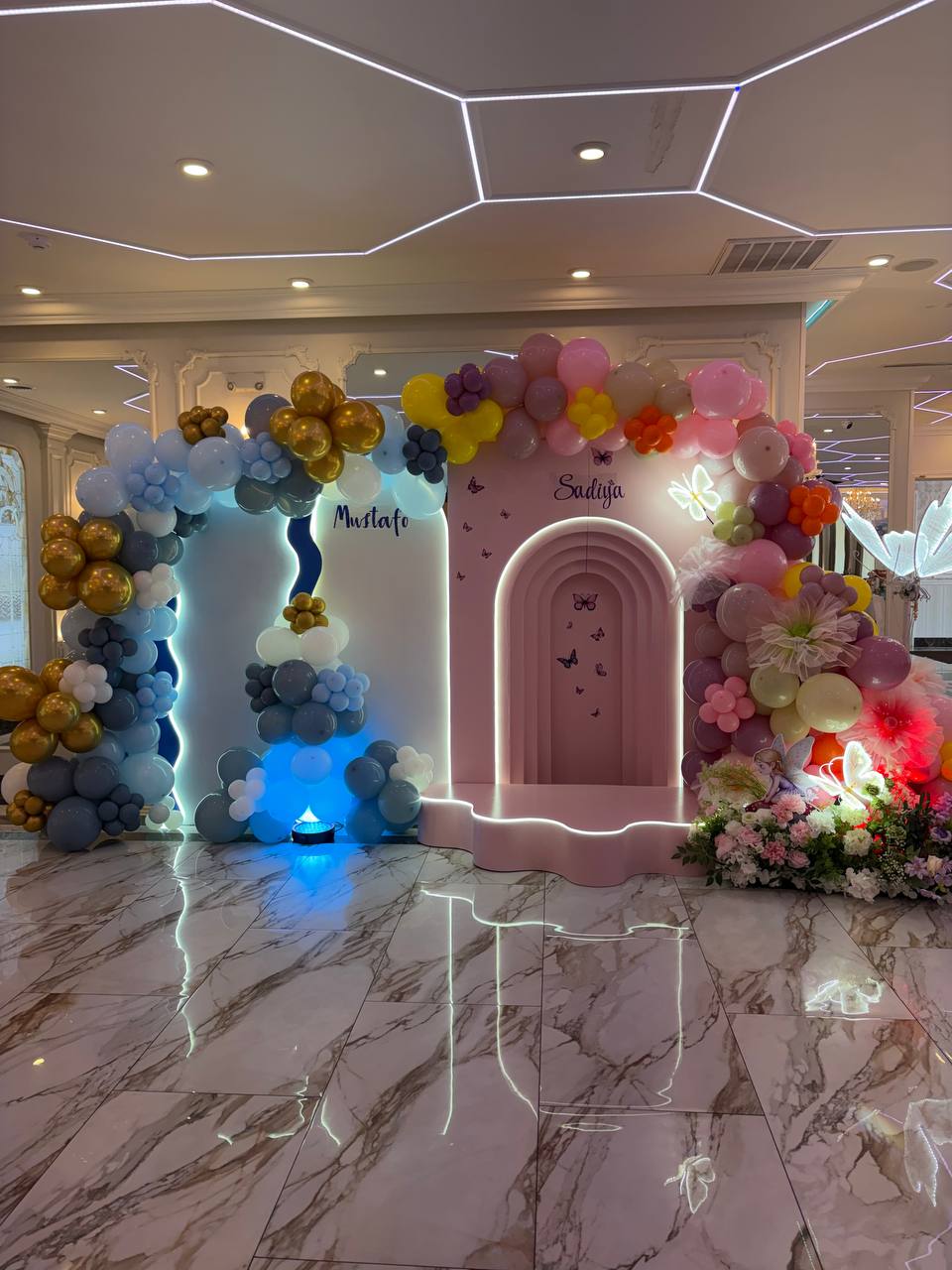 Two arched party backdrops side by side, one blue and gold and one pink, wrapped in balloon garlands and flowers for a twin birthday.