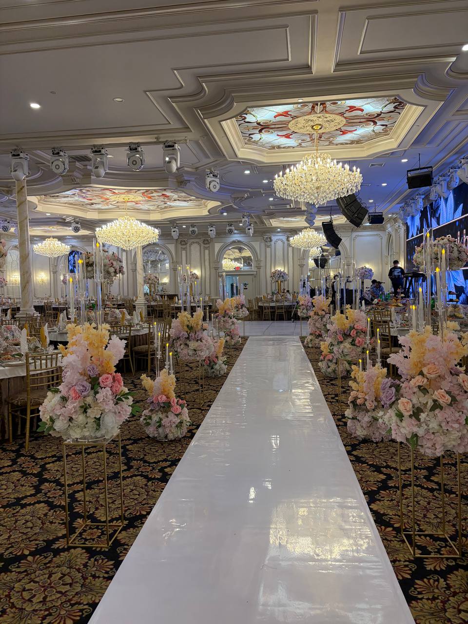 A ballroom aisle lined with tall blush, peach and lavender arrangements on gold stands beneath crystal chandeliers.