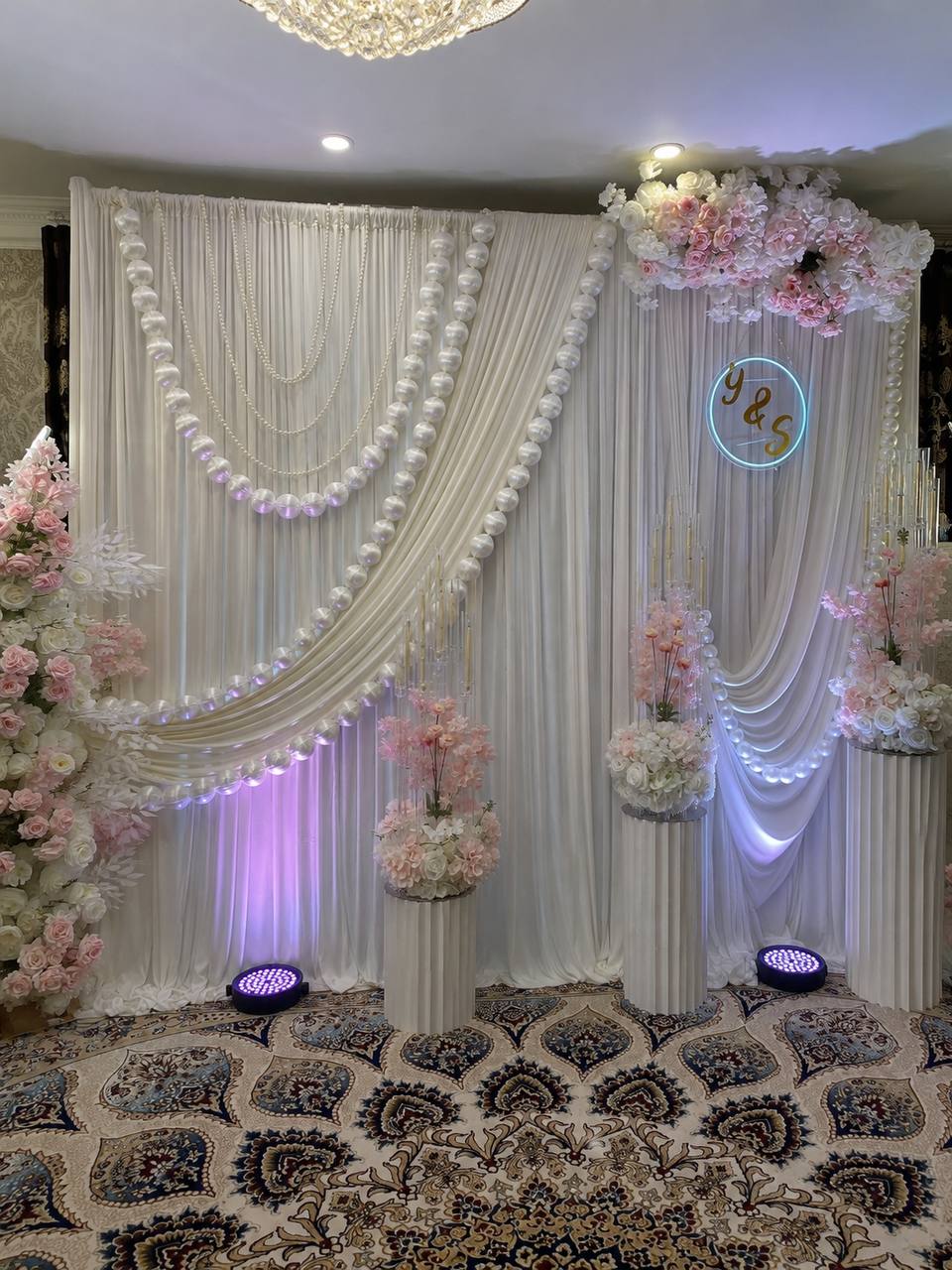 An ivory drape backdrop swagged with strands of large pearls, a Y & S neon monogram and pedestals of blush blossom.