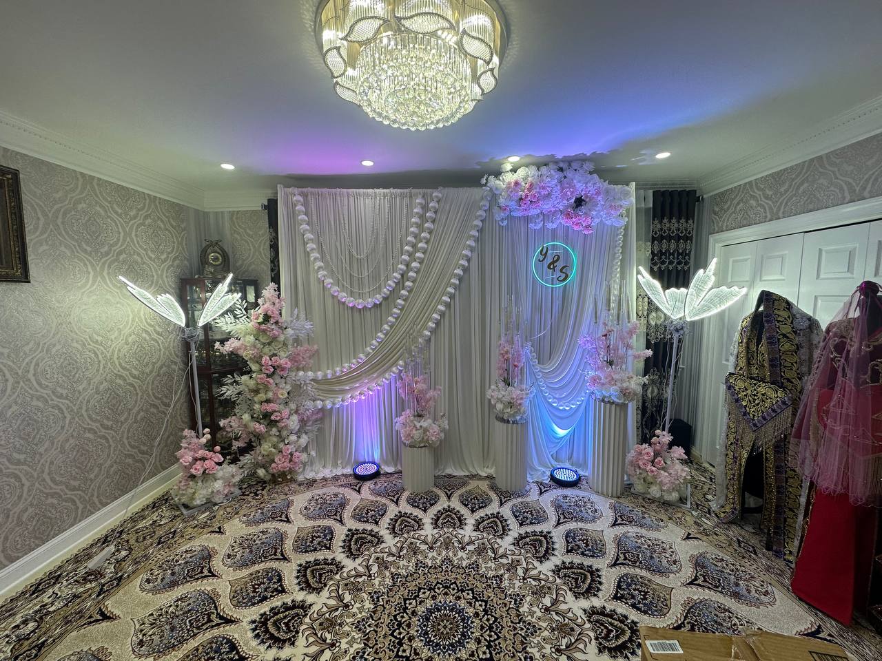 A living-room engagement setup with pearl-draped ivory panels, a Y & S neon monogram, blush blossom columns and lit wing sculptures.