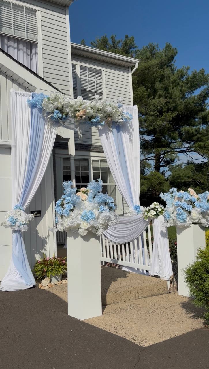 A home entrance dressed for a celebration with white drapery and clusters of powder-blue and white flowers on white columns.
