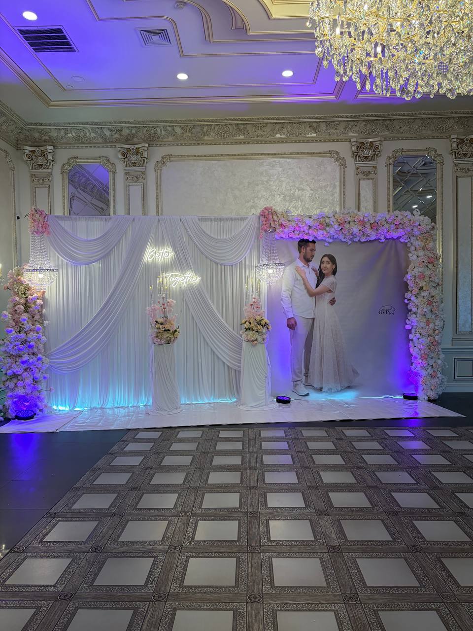 A white draped photo backdrop with a Better Together neon sign, crystal chandeliers and an arch of blush roses on a mirrored dance floor.