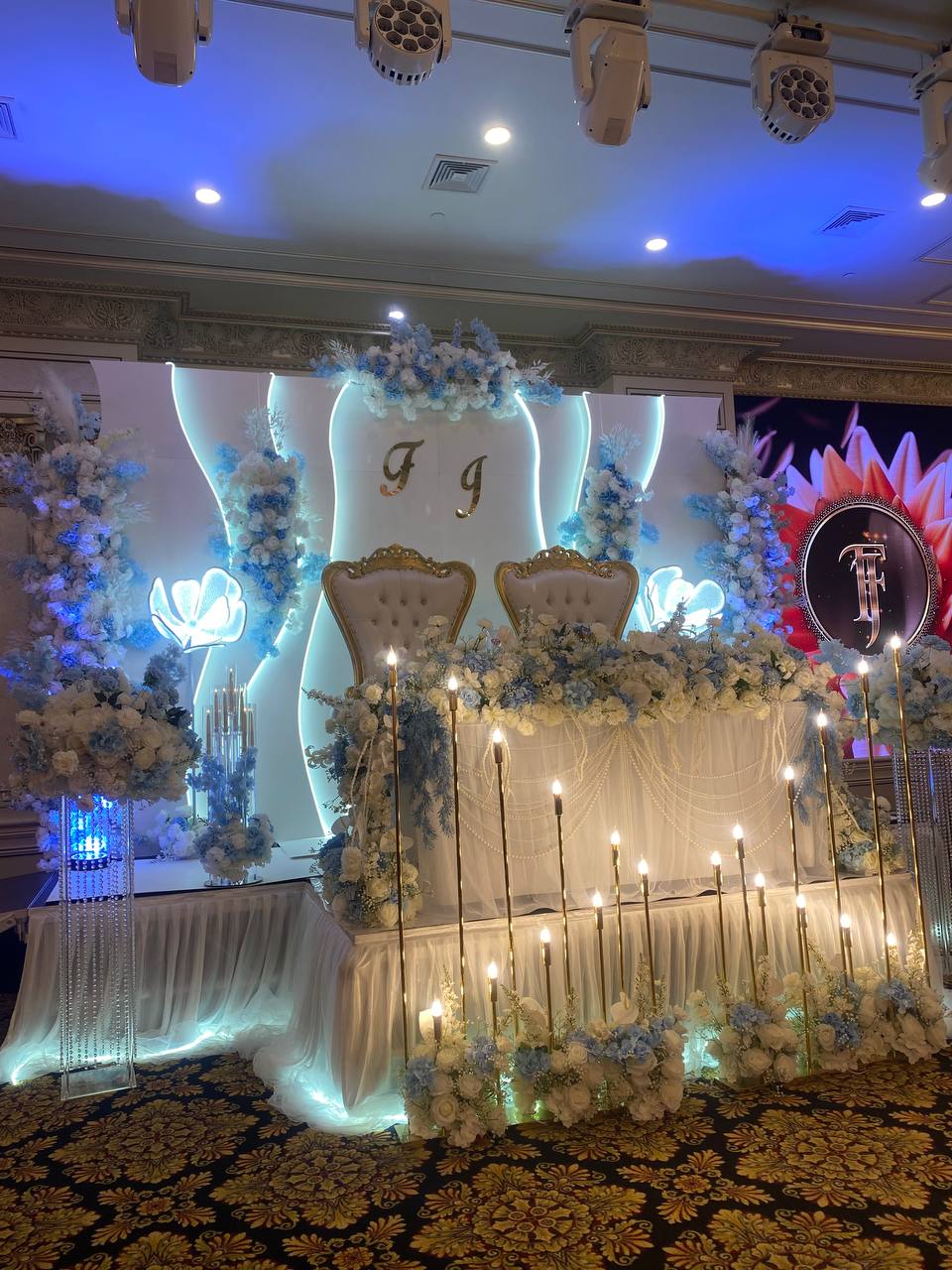 An angled view of a blue and ivory head table with gold thrones, a monogram medallion and rows of gold taper candles.