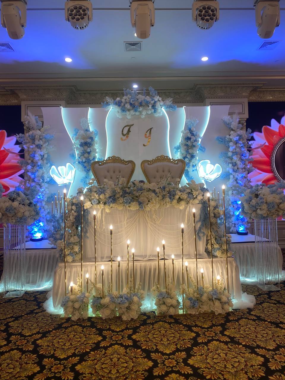 A wedding head table beneath a lit monogram wall, banked with blue and white florals and lined with tall gold taper candles.