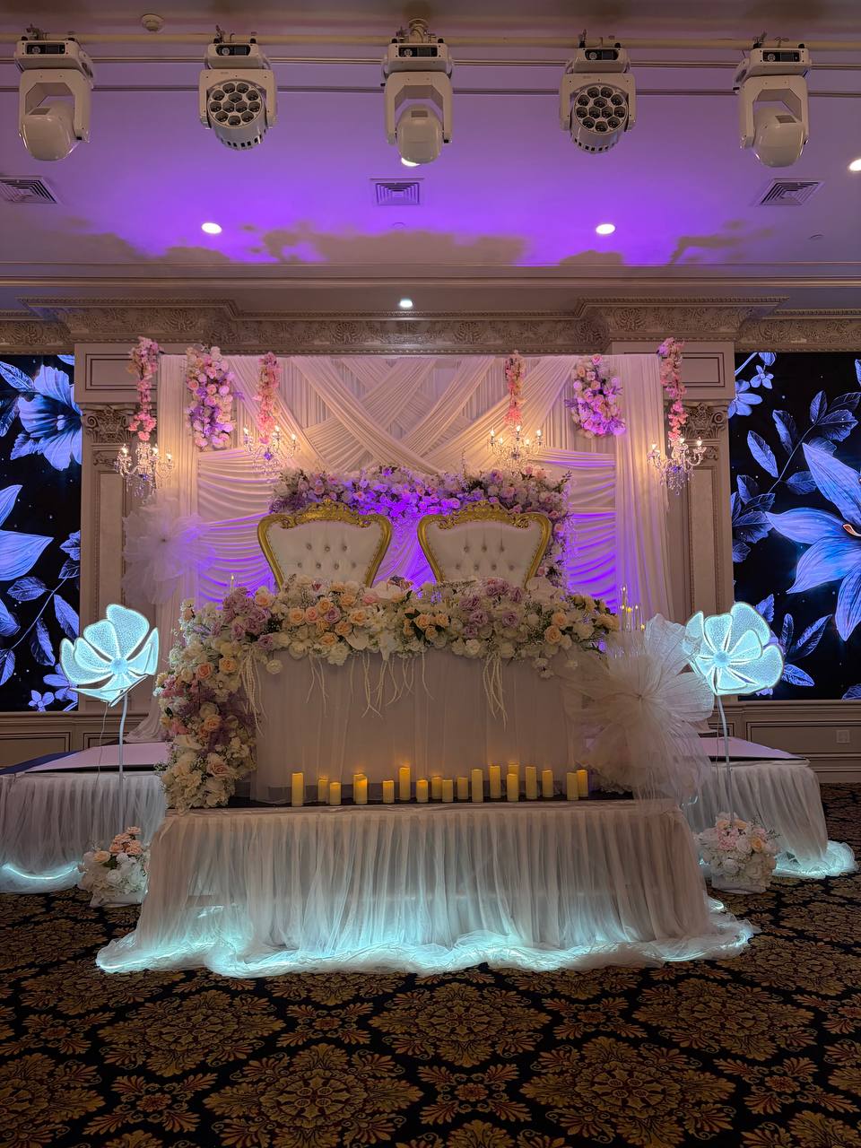 A sweetheart table with gold tufted thrones, a cascade of blush and ivory roses and a row of pillar candles under violet uplighting.